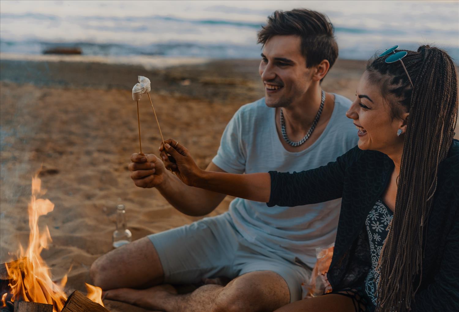 Couple enjoying a romantic evening on a Cornish beach, toasting marshmallows on an open fire with the sand and sea in the background