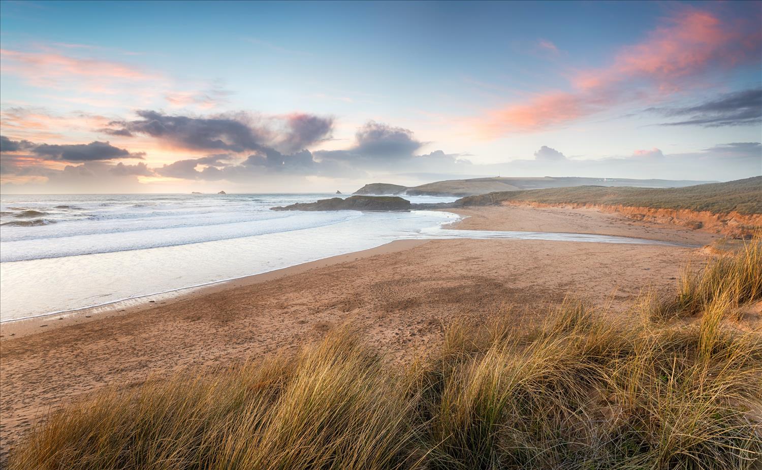 The sand dunes give way to sand on a deserted Constantine Bay beach, with gentle waves lit up pink as the sun sets