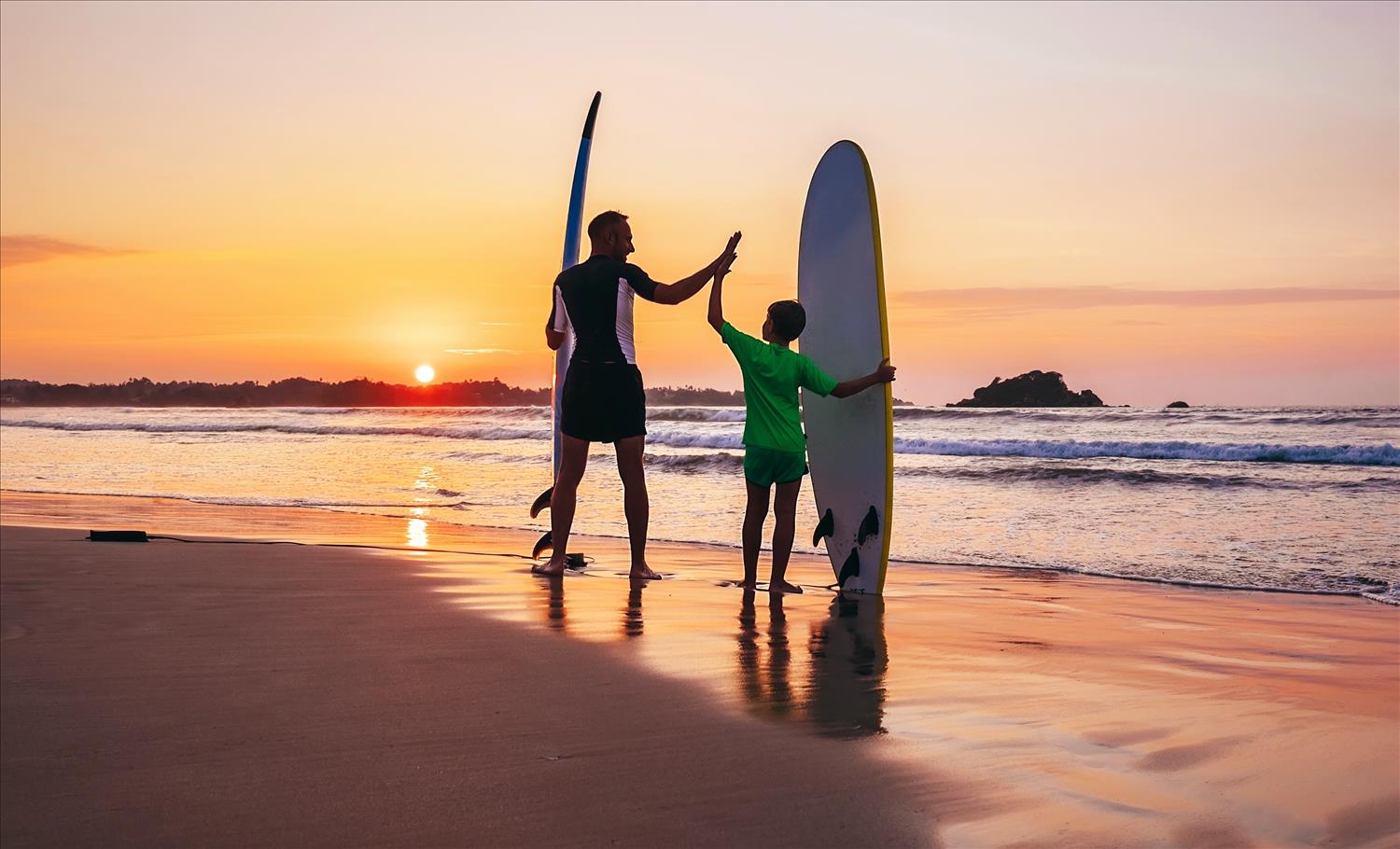 Father and son high-five as they stand in the sand with their surf-boards at their sides, watching the sun set