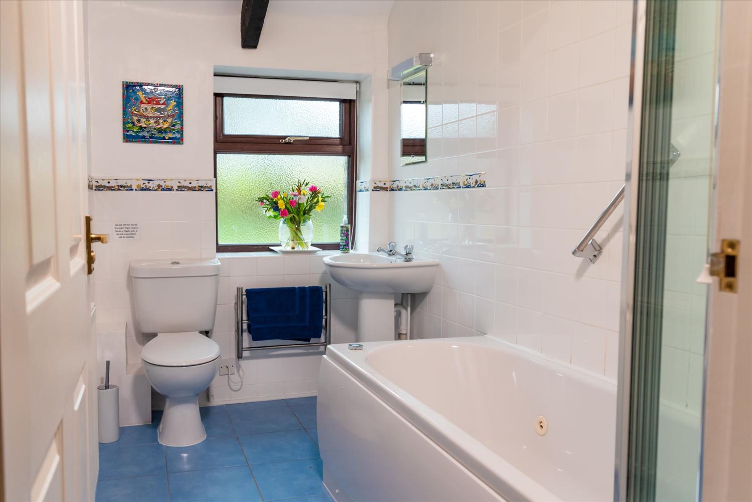 The bathroom at Polrunny Farm's Blackberry Cottage, with Whirlpool bath and flowers in the window and brightly coloured art on the wall.