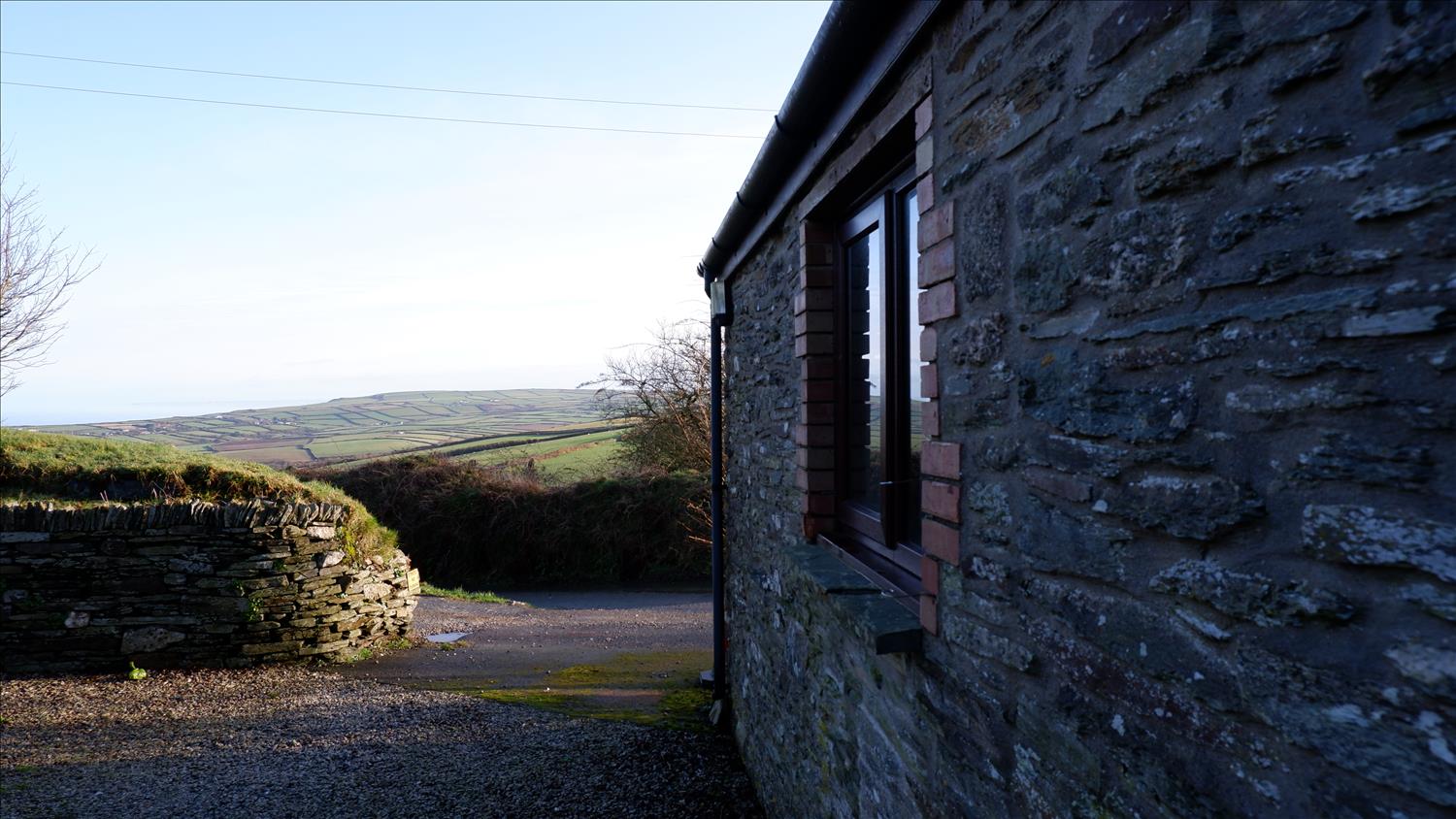 The stone wall of Polrunny Farm's Elderberry Cottage in the foreground, with stunning rural views beyond, as the hills roll down to the sea