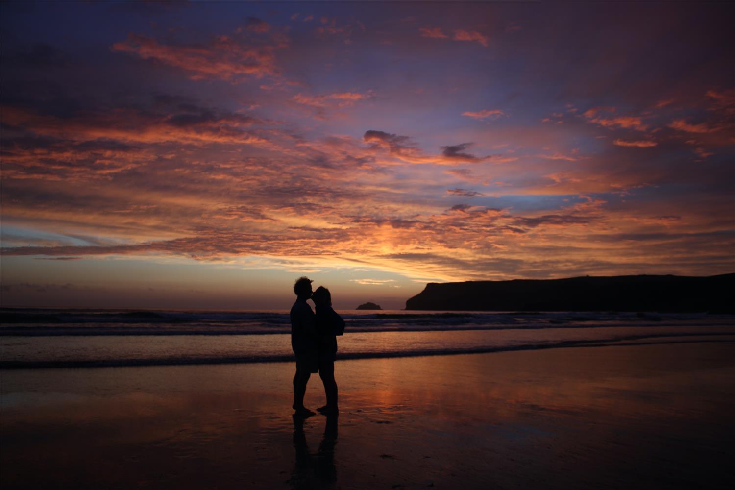 A couple kissing as they stand on the wet sand, with the sky lit up orange, pink and purple behind them as the last vestiges of light depart from Polzeath beach, North Cornwall