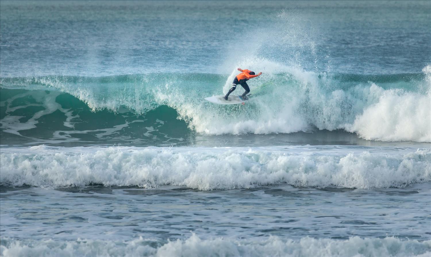A surfer in a bright orange top rides the waves at Fistral Beach, Newquay
